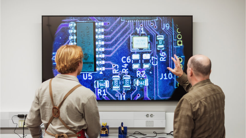 A student and faculty inspect a circuitboard under a microscope.